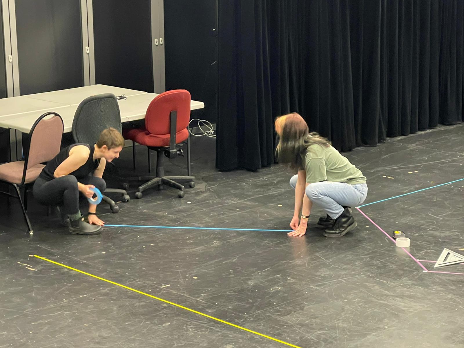 Image of two people in kneeling positions extending a line of tape accross the theater floor.