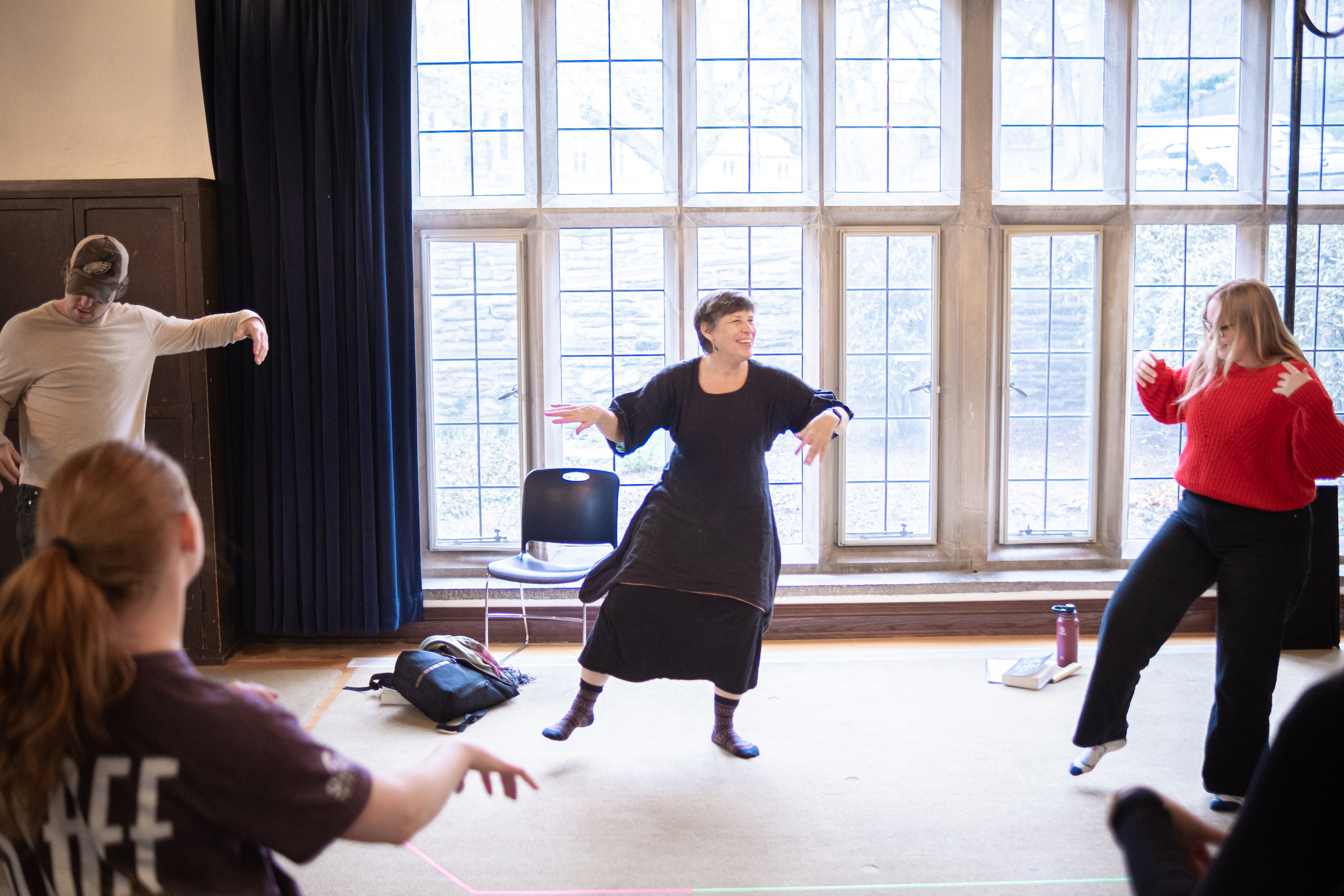 Image of Professor Catherine Slusar leading a group of students in an acting exercise. She is in the center of the image with one foot held off of the ground and arms in motion. Three students are visible in the frame, one to each side of Professor Slusar and one in the foreground.