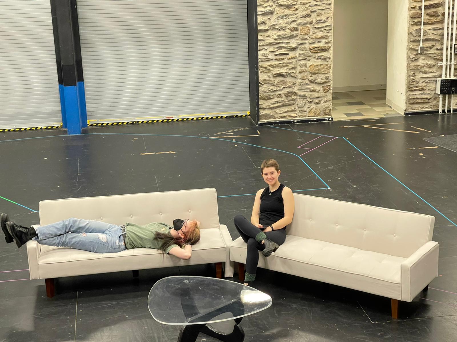 Image of two people smiling and sitting on a couch on a theater set.