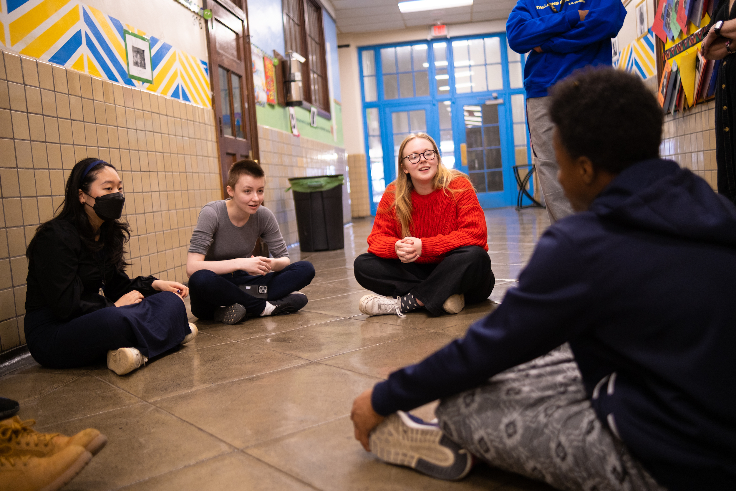 Three young-adults sit on the floor and speak to two other pictured individuals, one is sitting and the other standing.