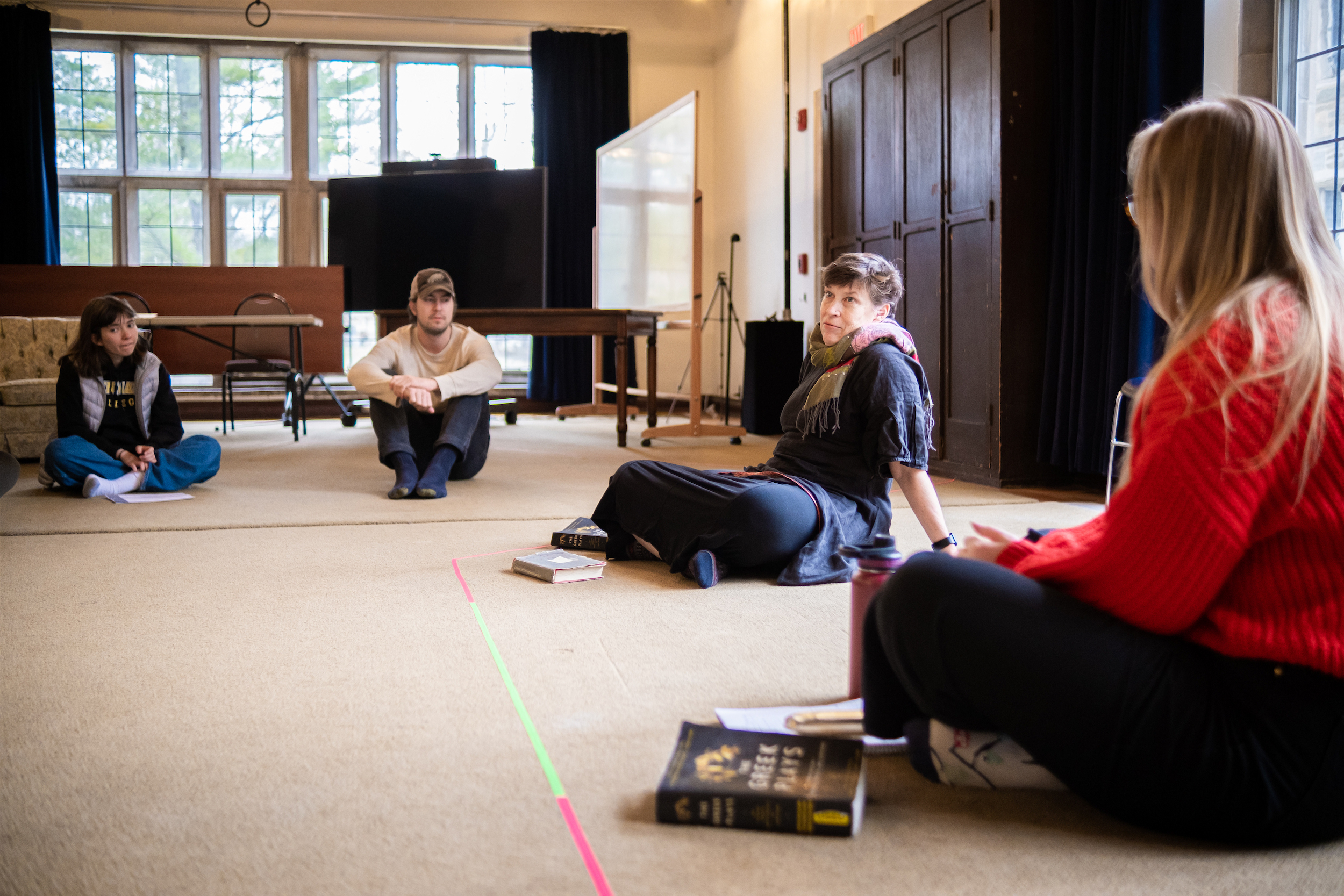 Four people are seated on the floor, some with a book or notebook in front of them. One person speaks as the rest listen.