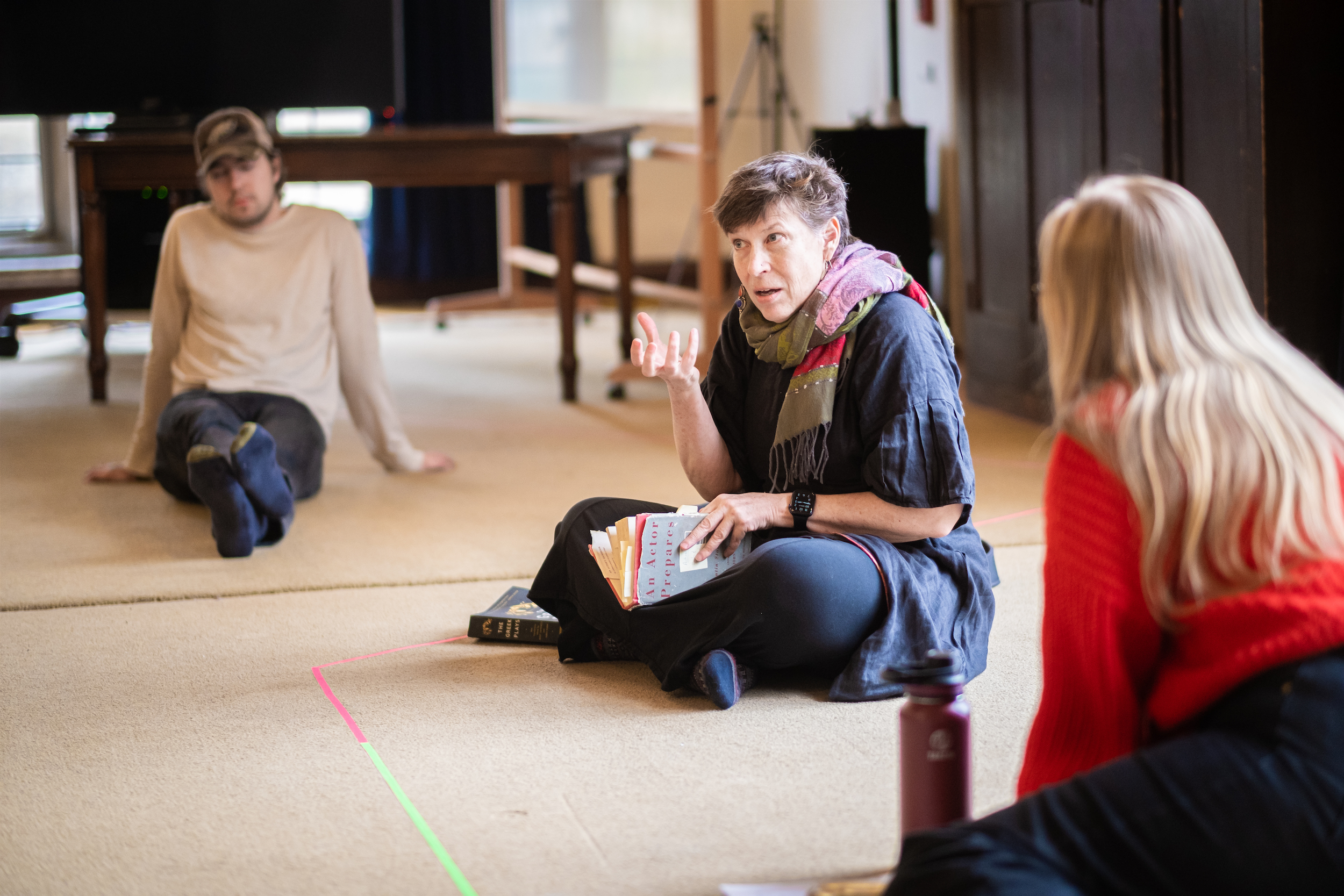 Three people are seated on the floor. One reads aloud from a book and gestures, the others listen.