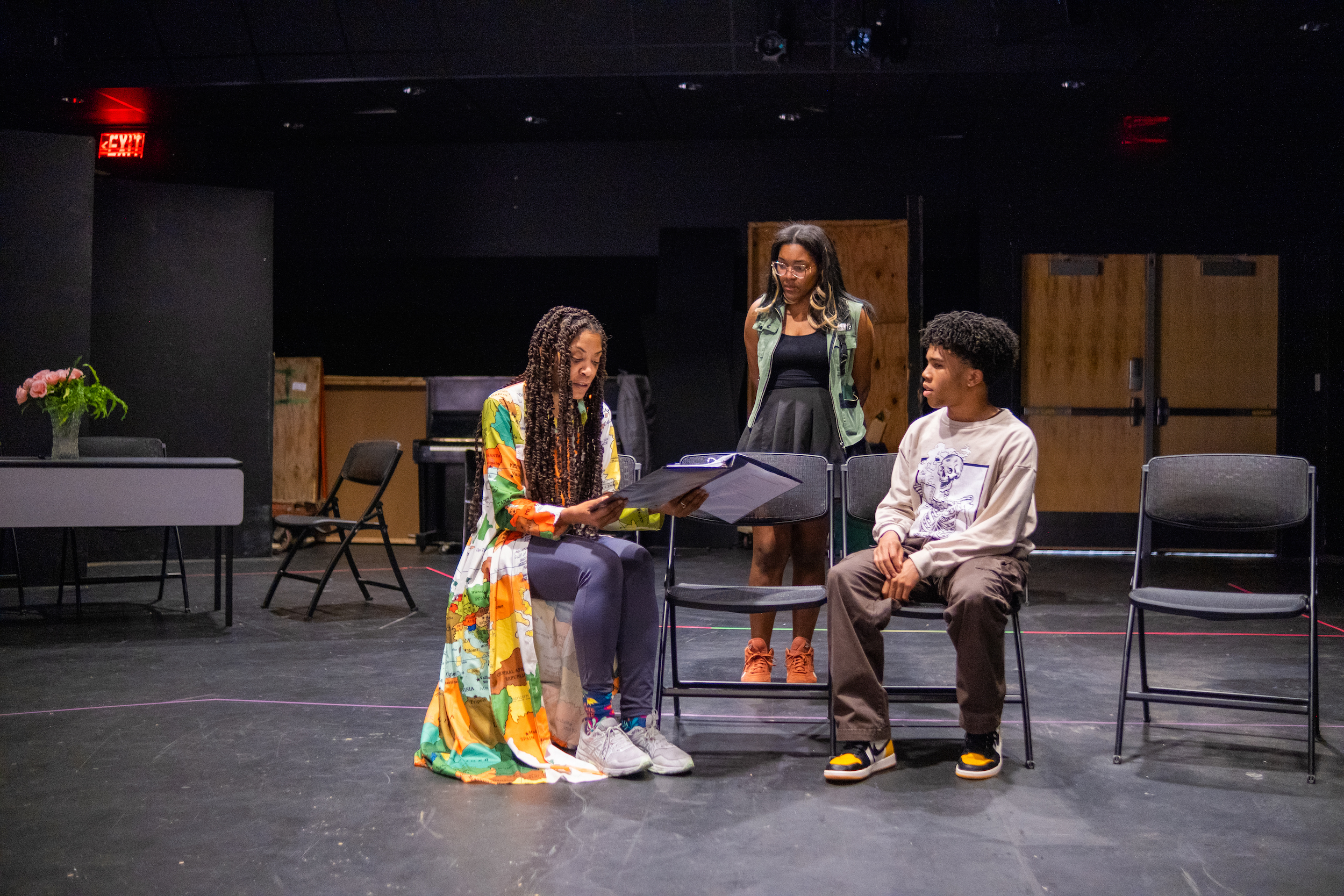 Three figures in a rehearsal space. Two are seated, one holding a script. The third person stands behind them.