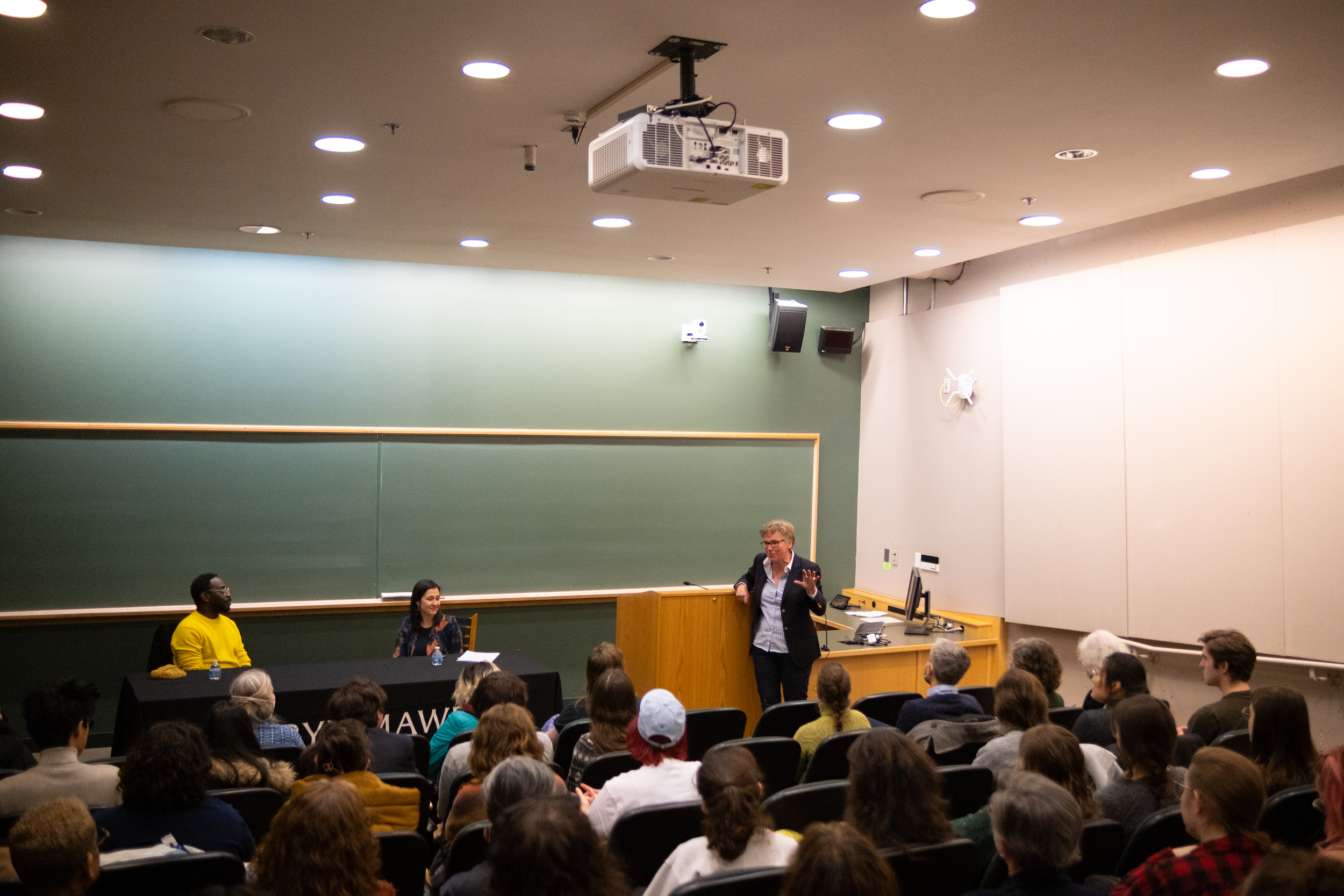 Three people at the front of a lecture hall before an audience. One is standing and speaking. The two others are seated at a table and listen to the speaker.