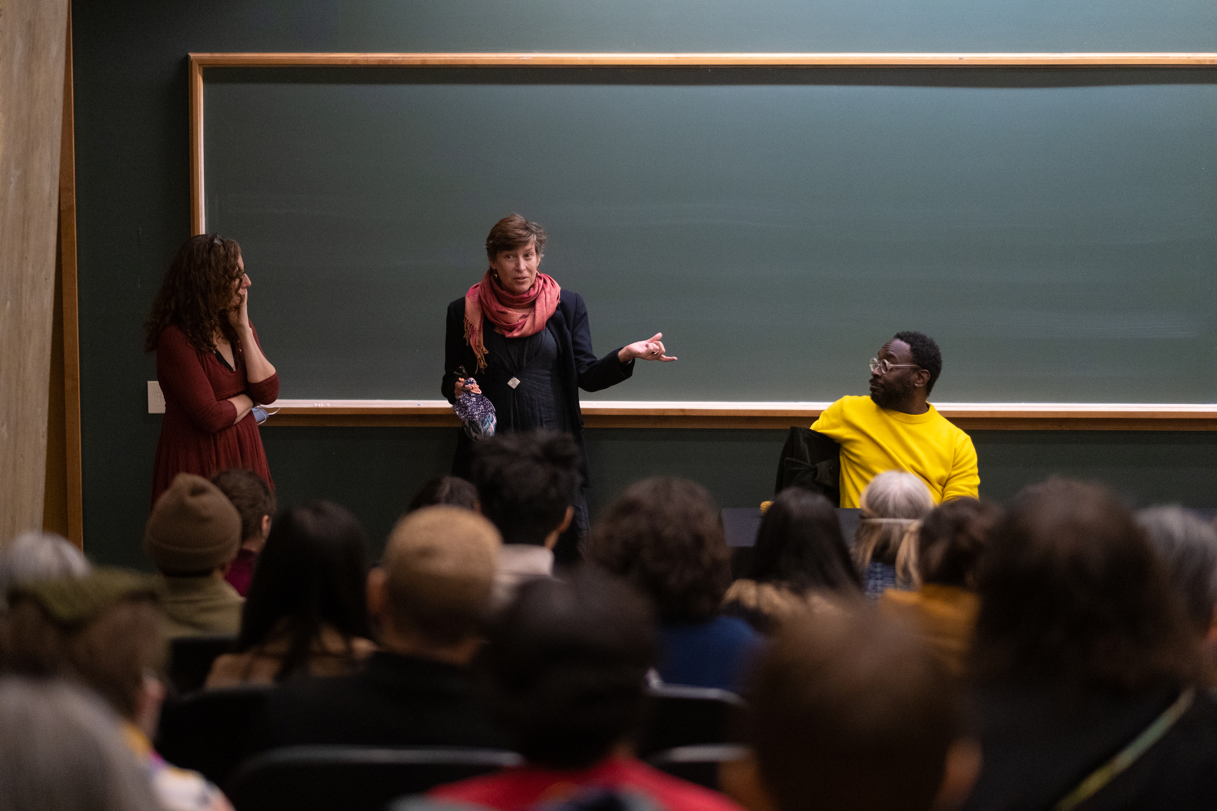 Three people at the front of a lecture hall before and audience. Two are standing, one contemplating with chin in hand as the other speaks. The third person is seated at the end of a table and listens.