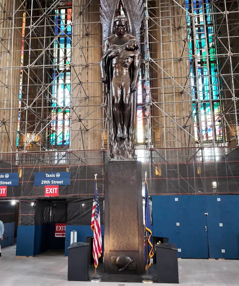 A photograph of a bronze sculpture titled Pennsylvania Railroad War Memorial by Walker Kirtland Hancock. It depicts an angel holding a lifeless man by his arms above flames. The sculpture is placed on a granite pedestal with the names of Pennsylvania Railroad employees who died in World War II.