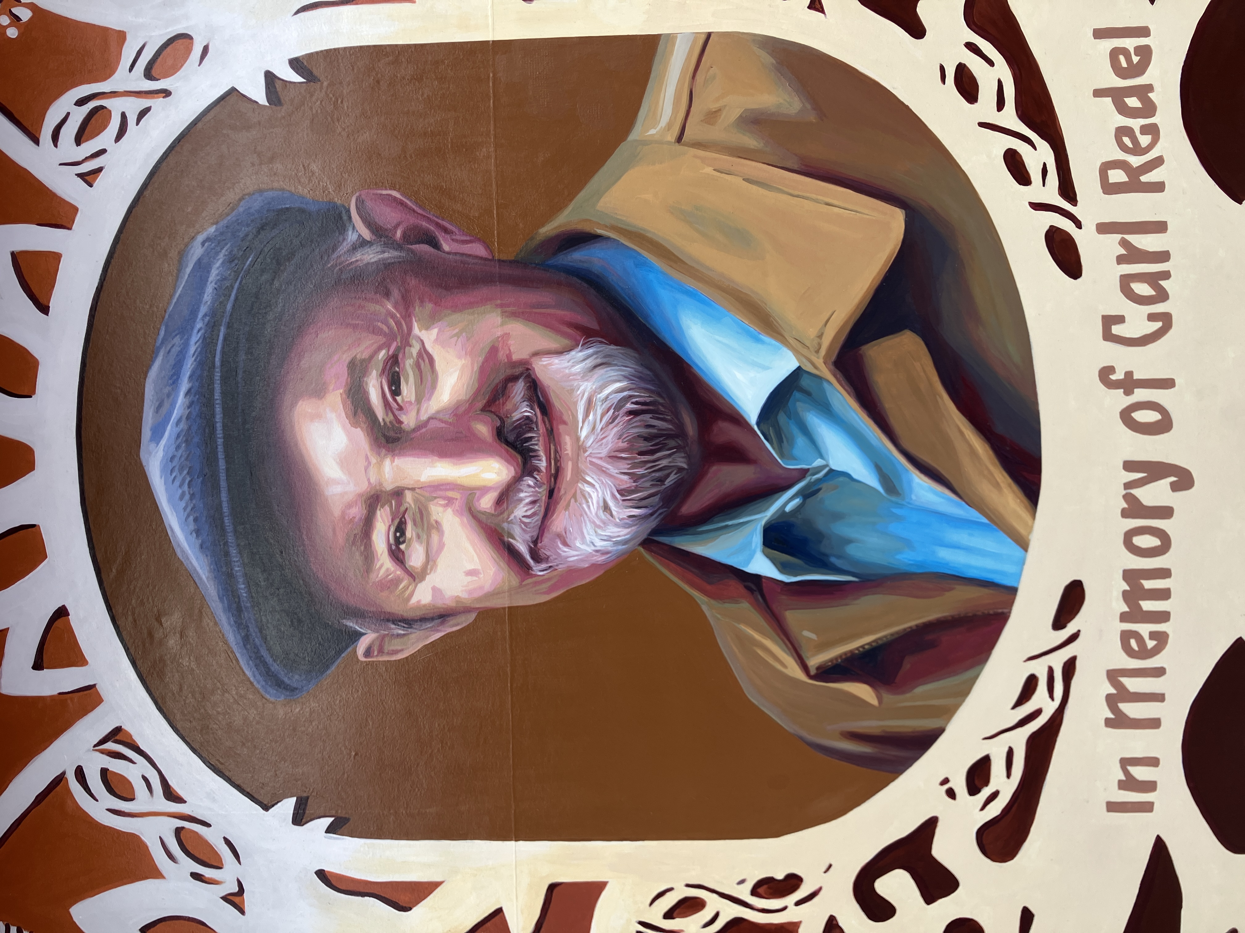 A mural on a brick wall showing a man smiling at the viewers, with a frame around him that resembles a white paper-cut pattern on a red background.