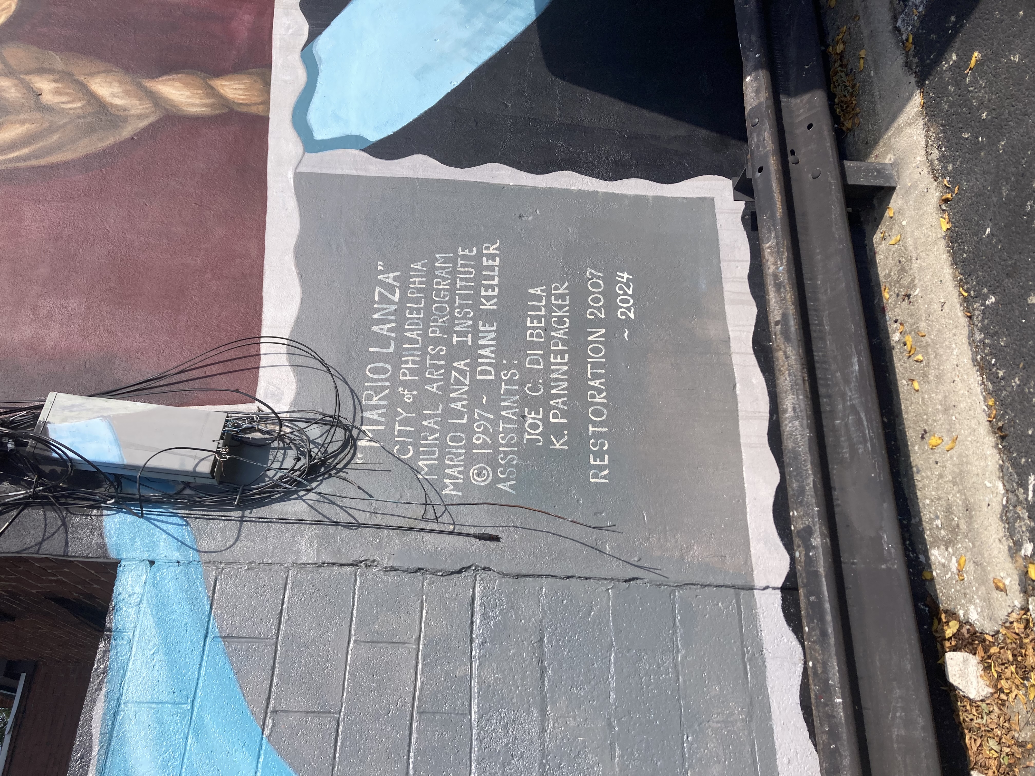 A list of contributors after the mural’s title, in white text on gray background. City of Philadelphia Mural Arts Program, Mario Lanza Institute, Diane Keller (assistants Joe C. Di Bella, K. Pannepacker). Lists mural creation as 1997 and restorations as 2007 and 2024