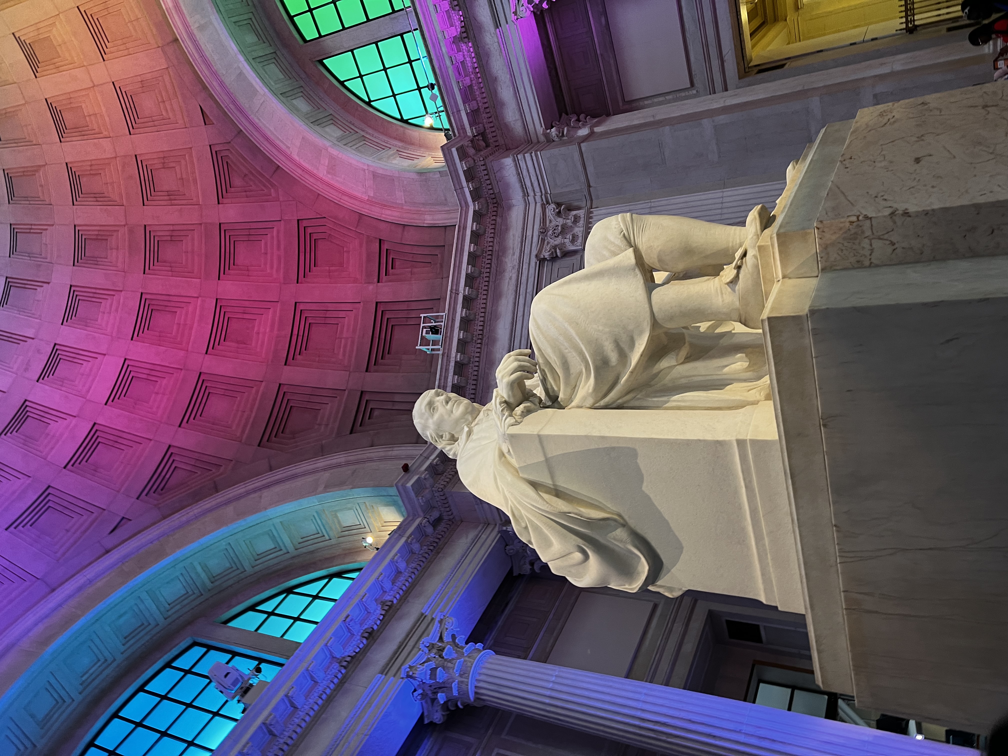 (Placeholder image) Low angle shot of sculpture of Benjamin Franklin seated. Franklin institute rotunda can be seen in the background illuminated by colorful lights.