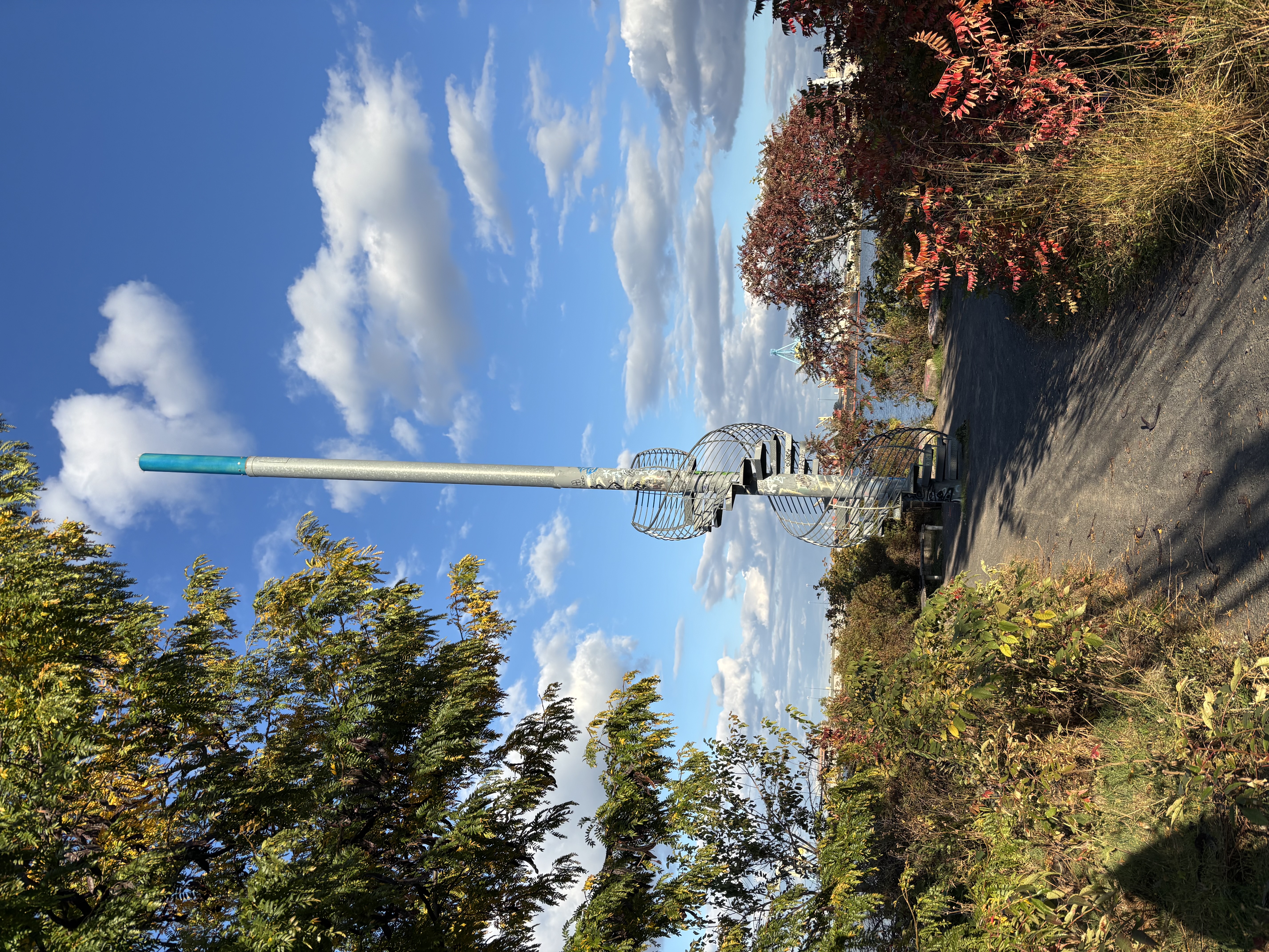 The "Land Buoy" is a spiral staircase that encircles a tall pole with a blue section at the top. Most of the background is a partially cloudy sky, and there is are bushes and trees on either side of the path to the spiral staircase. 