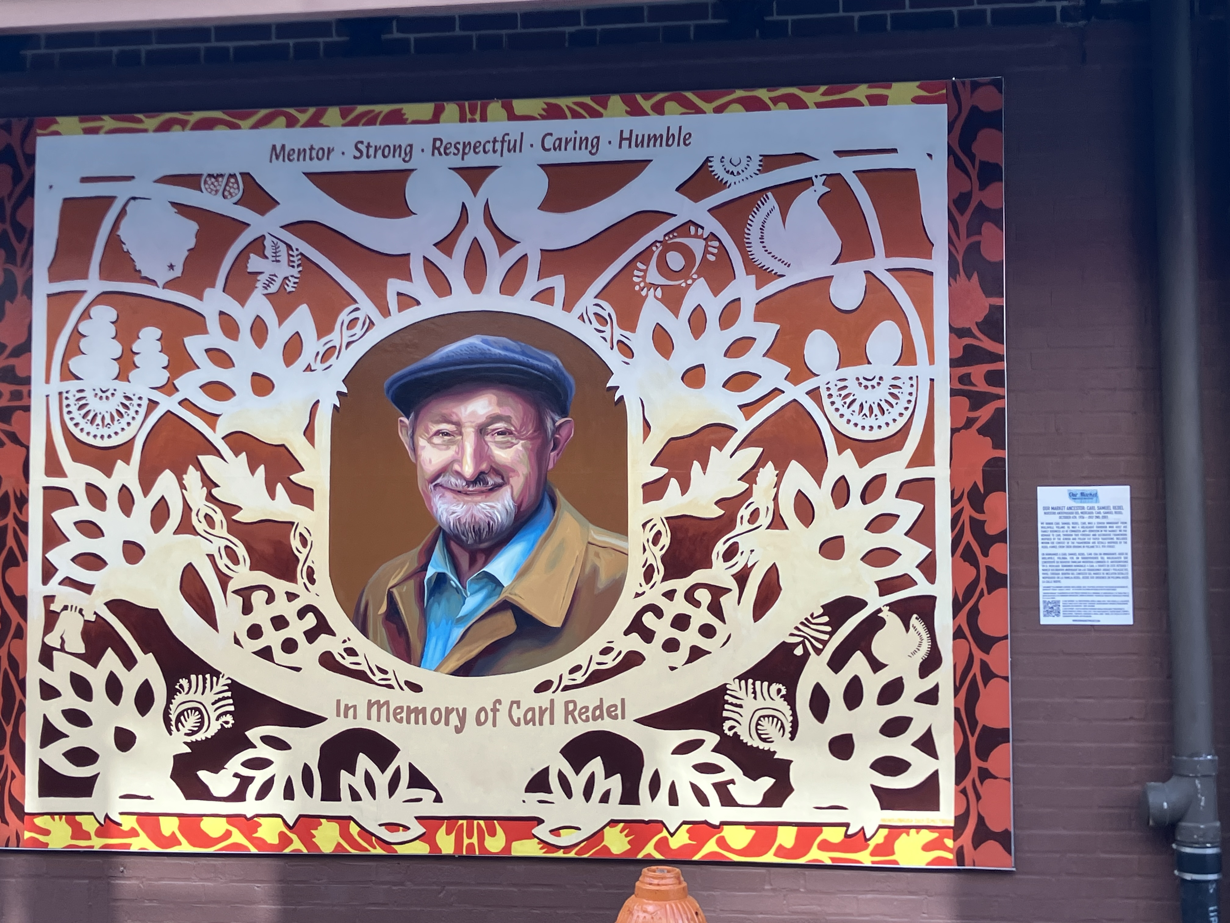 A mural of an elderly man smiling at the viewer, with a frame that looks like cut paper painted around him. Below him text reads "In Memory of Carl Redel," and above him text reads "Mentor - Strong - Respectful - Caring - Humble."