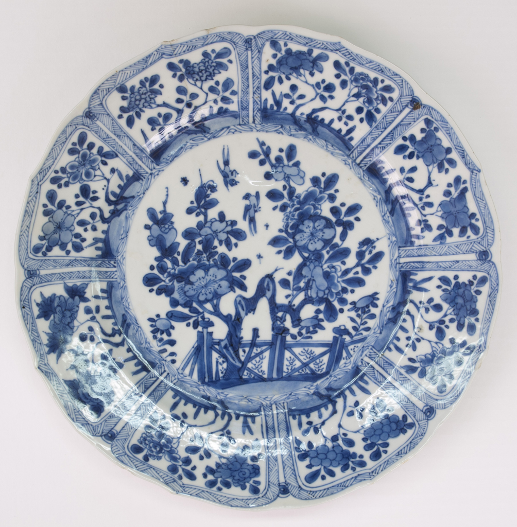 Photo of a 20th century Caton ware plate. The ceramic plate is white with dark blue detailing depicting a trees and a fence.